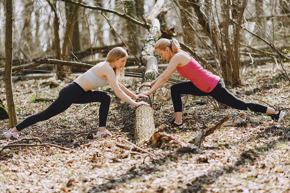 Zwei Frauen stützen sich beim Sport im Wald auf einen Baumstamm, um sich zu dehnen.